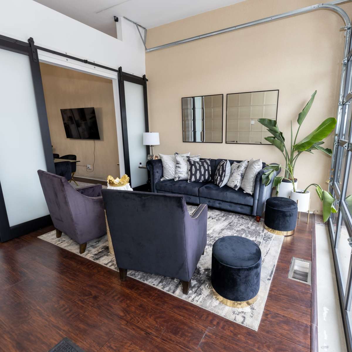 A modern living space features a dark sofa, two armchairs, and decorative pillows on a patterned rug, with a large window providing natural light.