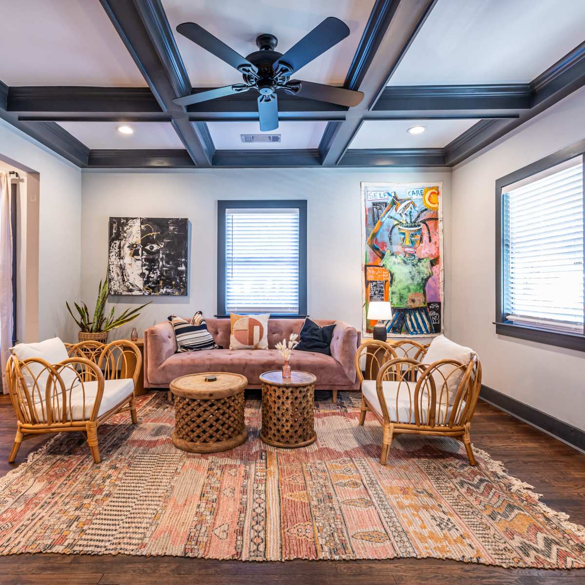 A modern living room with a coffered ceiling, featuring two rattan chairs, a pink sofa, a round wooden coffee table, and colorful artwork on the walls.