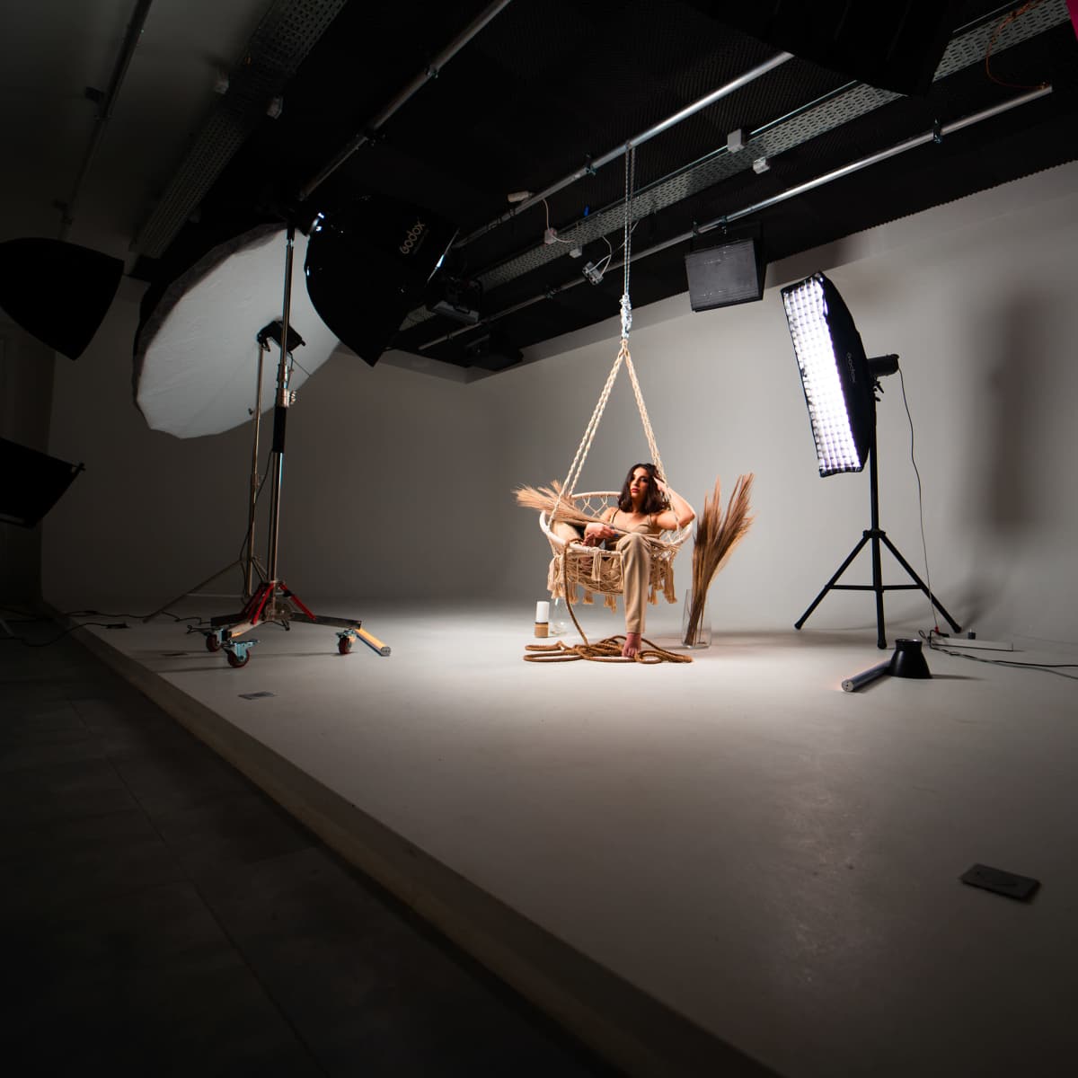 A person sits on a swing suspended from the ceiling in a well-lit studio with various lighting equipment.