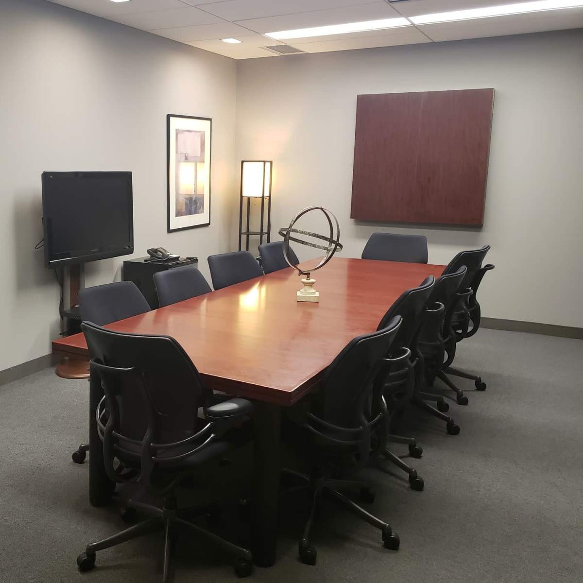 A conference room with a large wooden table surrounded by chairs, a television on the wall, and framed artwork.