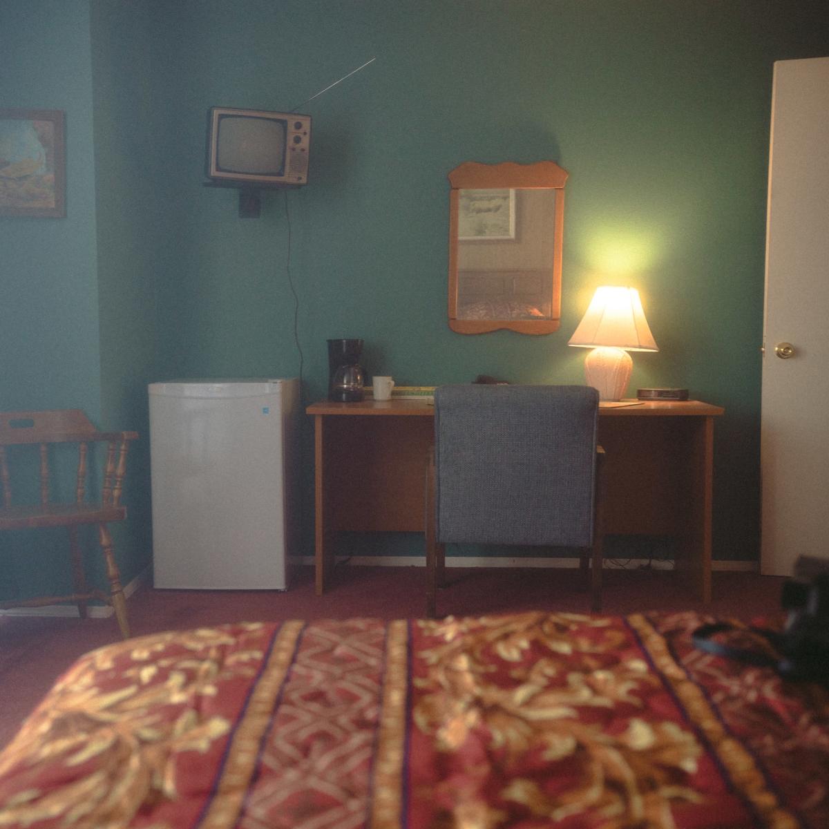 A small hotel room features a chair, a desk with a mirror, a coffee maker, a mini-fridge, an old television mounted on the wall, and a door marked with the number four.
