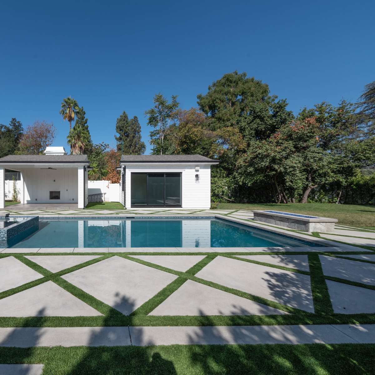 A modern backyard featuring a swimming pool surrounded by geometric stone pathways and landscaped greenery, with a shaded seating area and poolside structures.