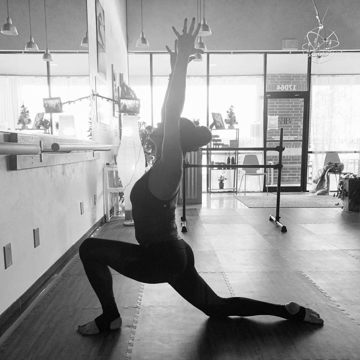 A person performs a yoga pose in a brightly lit studio, stretching one arm overhead while kneeling with the other leg extended forward.