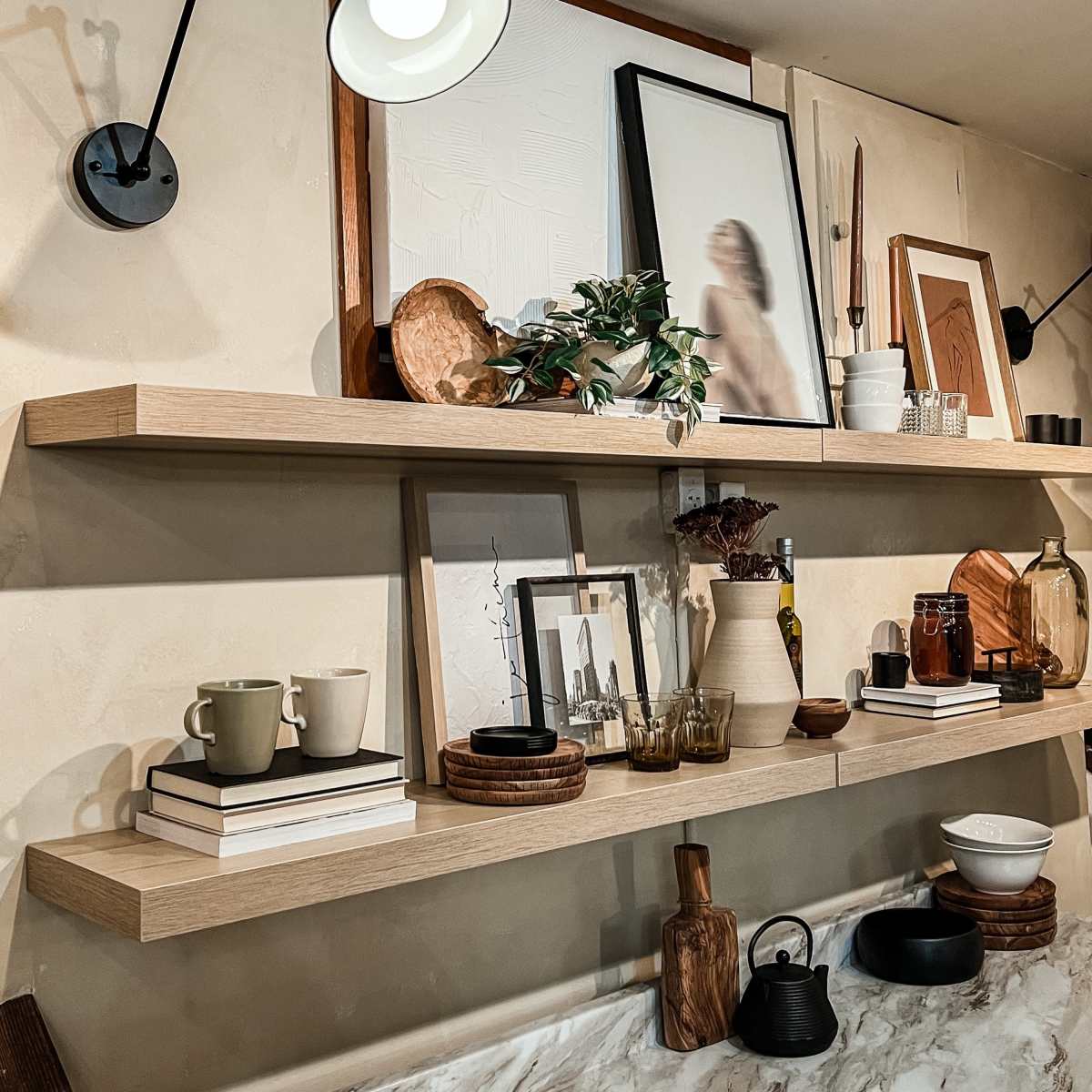 A wall-mounted shelf with various decorative items, including framed pictures, ceramic mugs, glassware, and wooden bowls, illuminated by a wall lamp.