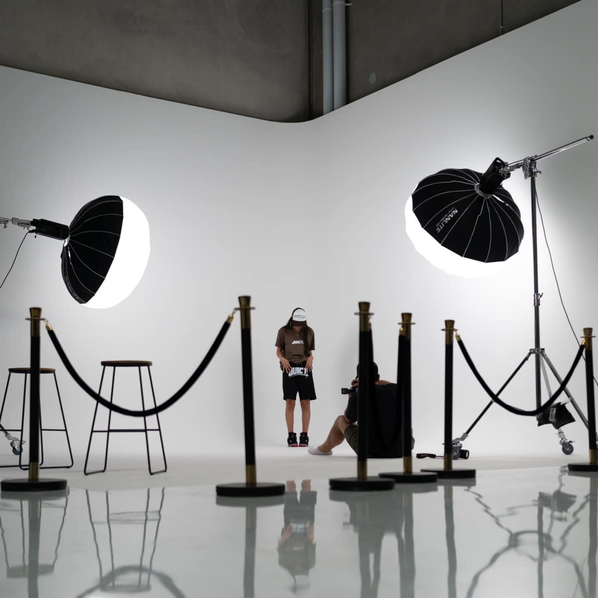 A photoshoot setup features a model and a photographer in a studio with two large light sources and a roped-off area.