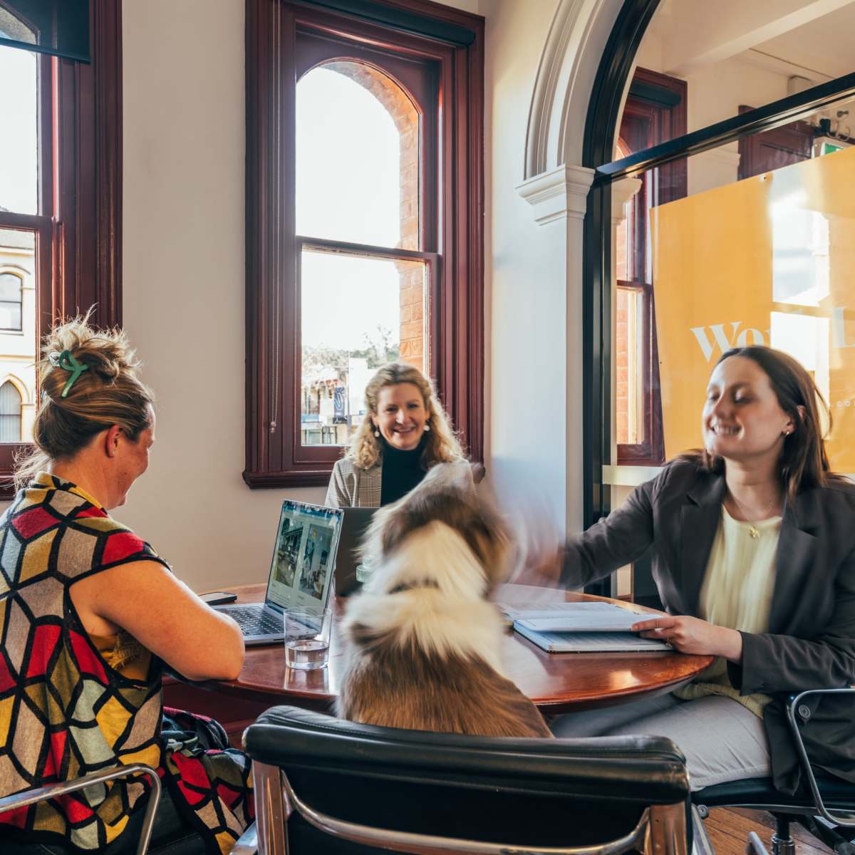 Two women are sitting at a table in a cafe, one with a laptop, while a dog plays nearby, and another woman is smiling in the background near the window.