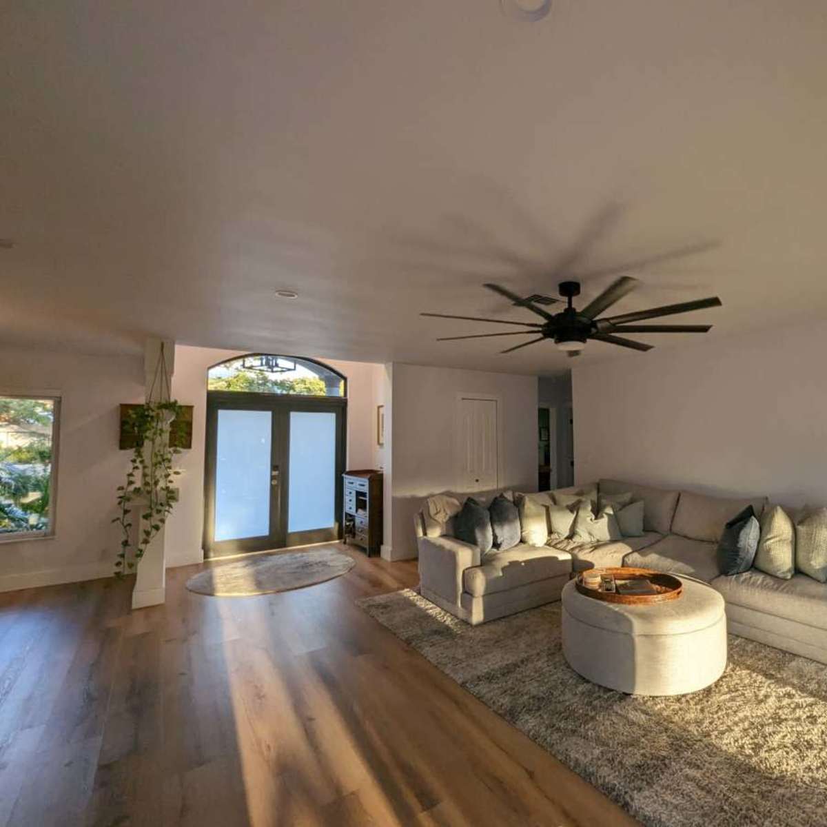 A spacious living room features a light-colored sectional sofa, a circular coffee table, and a large ceiling fan, with natural light streaming in through a large front door.