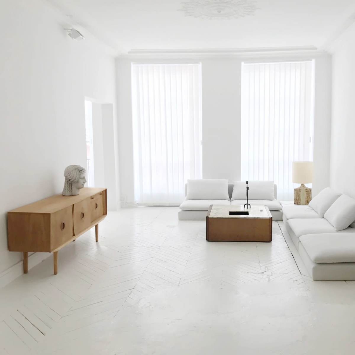 A minimalist living room with white walls, a light-colored wooden cabinet, a coffee table, and a sectional sofa arranged around a central space.