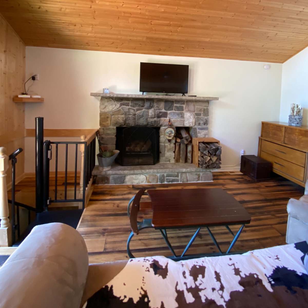A living room with a stone fireplace, a flat-screen TV mounted above it, and wooden flooring, along with a staircase and a wooden dresser.