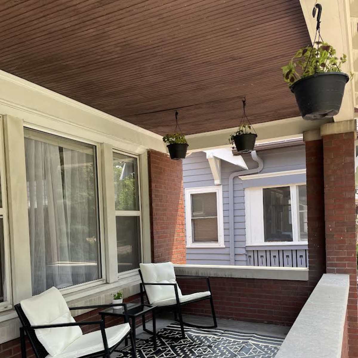 A covered porch with two white chairs and a small table on a patterned rug, surrounded by brick walls and adorned with hanging potted plants.