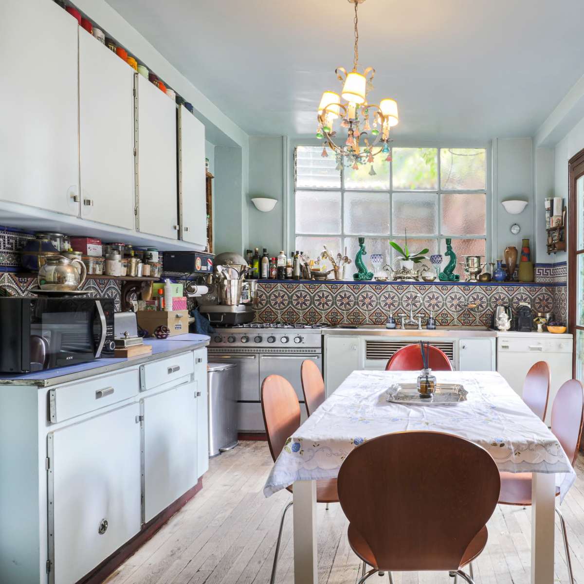 A kitchen features a central table surrounded by chairs, with cabinets and appliances along the walls and large glass doors leading to a garden.