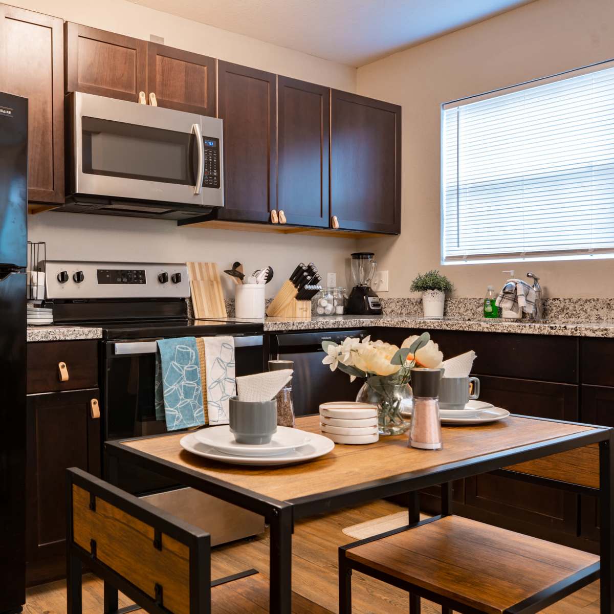 A modern kitchen features dark wooden cabinets, a black refrigerator, stainless steel appliances, and a dining table set with dishes and flowers.