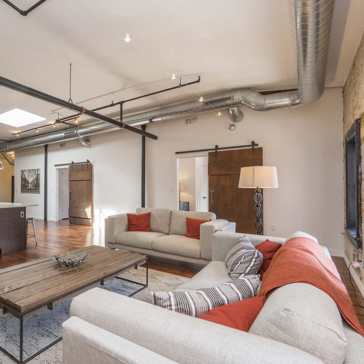 A spacious, modern living area with a kitchen to the left, featuring hardwood floors, industrial piping, and large sliding barn doors.
