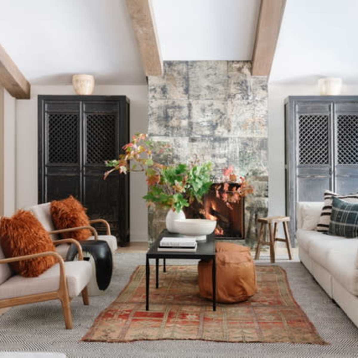 The living room features a stone fireplace with a floral arrangement on a coffee table, flanked by two dark cabinets, cozy seating, and a patterned area rug.