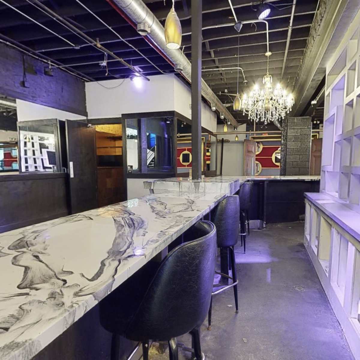 A modern bar area with a long, decorated countertop, high stools, and a chandelier overhead in a dimly lit space.
