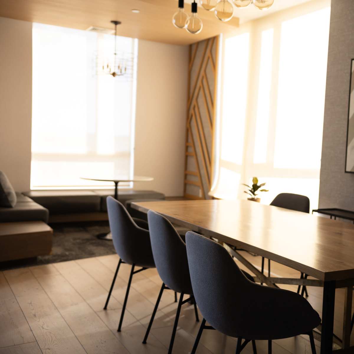 A modern meeting room features a large wooden table surrounded by upholstered chairs, with a wall art piece and sunlight streaming through large windows.