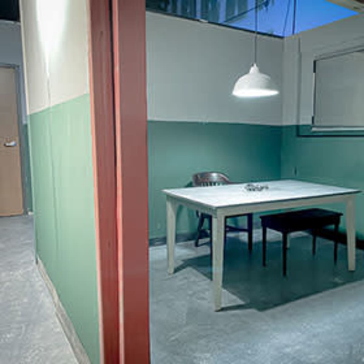 A sparse room featuring a table with two chairs, a filing cabinet, and a single hanging light fixture.