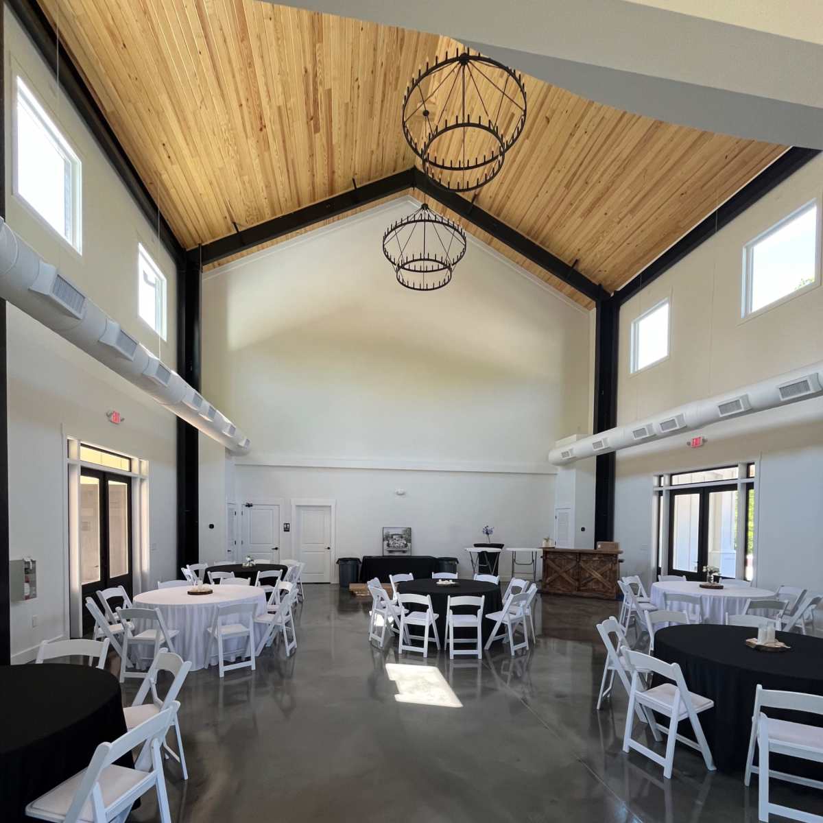An indoor event space with round tables covered in black and white tablecloths, set beneath a high wooden ceiling with industrial-style light fixtures.