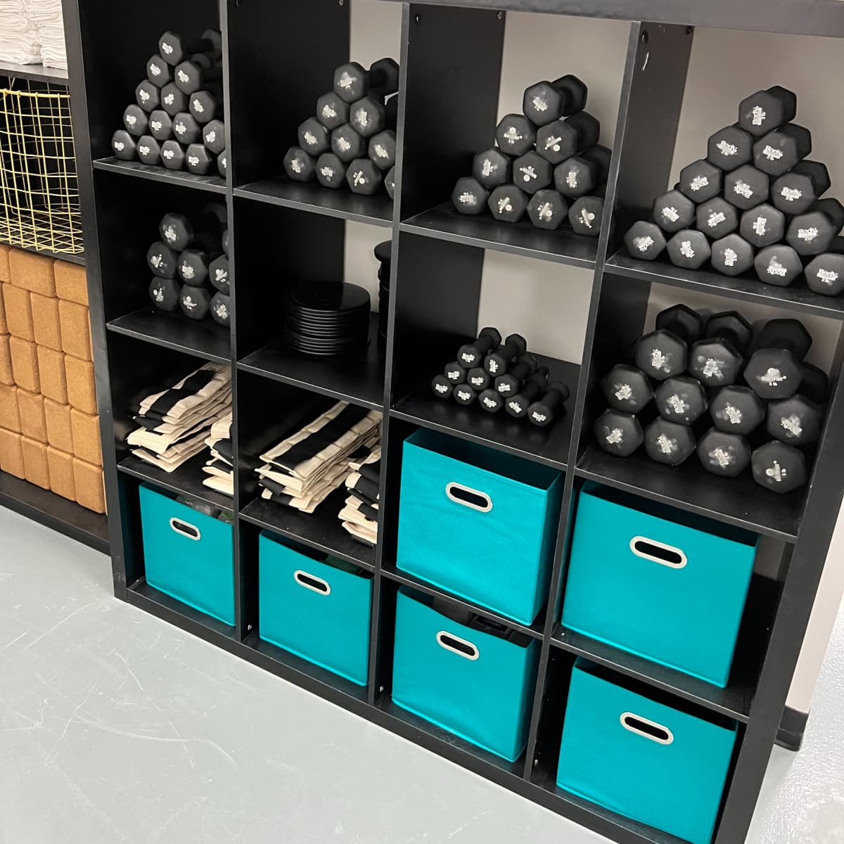 A black shelving unit contains neatly organized black dumbbells, weight plates, and storage bins in a workout space.