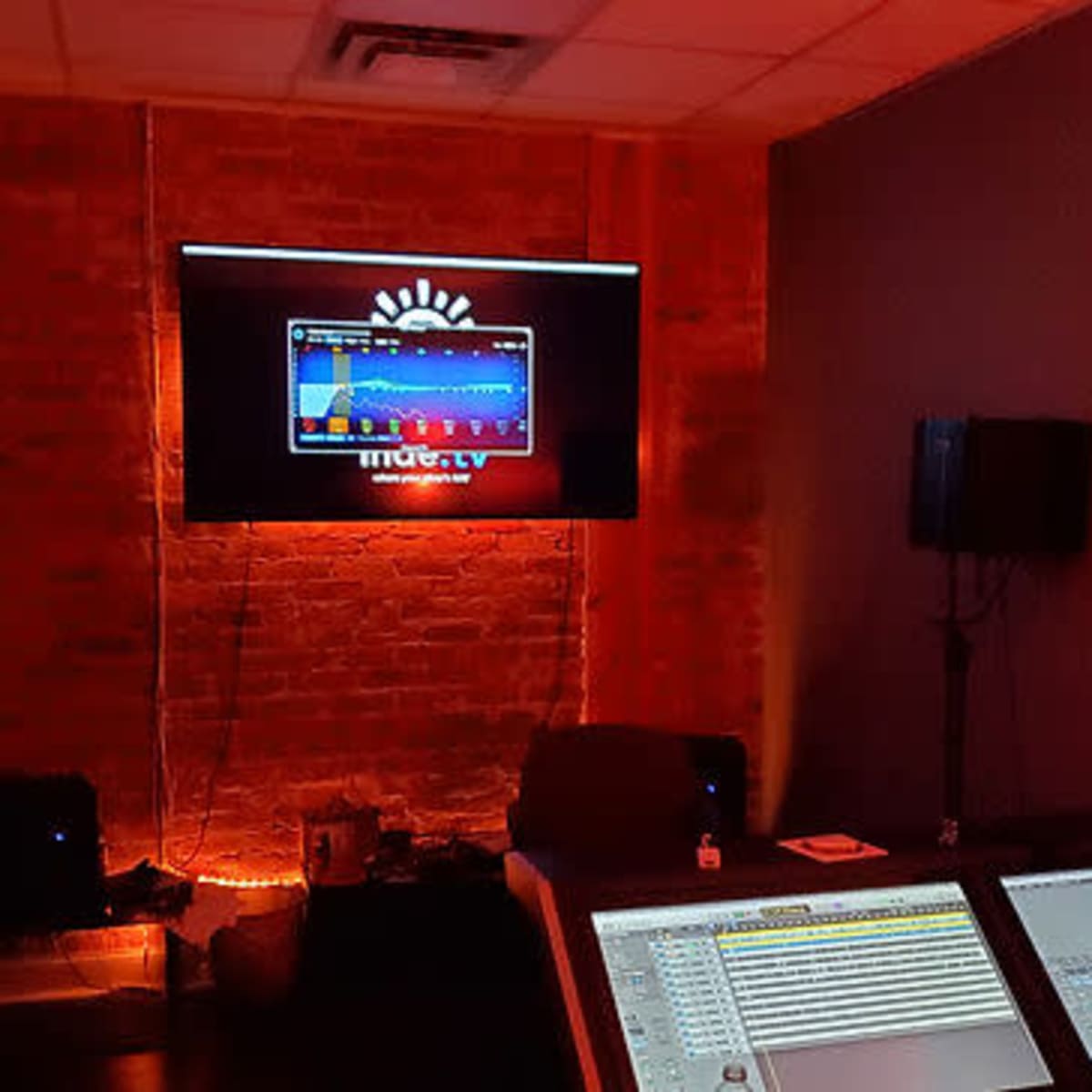 A person is seated in a dimly lit room, focused on a sound mixing console with a large screen displaying audio wave patterns in the background.