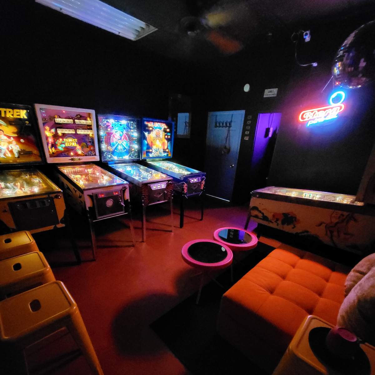 A dimly lit room featuring several pinball machines, neon lighting, and colorful furniture, including an orange couch and yellow stools.
