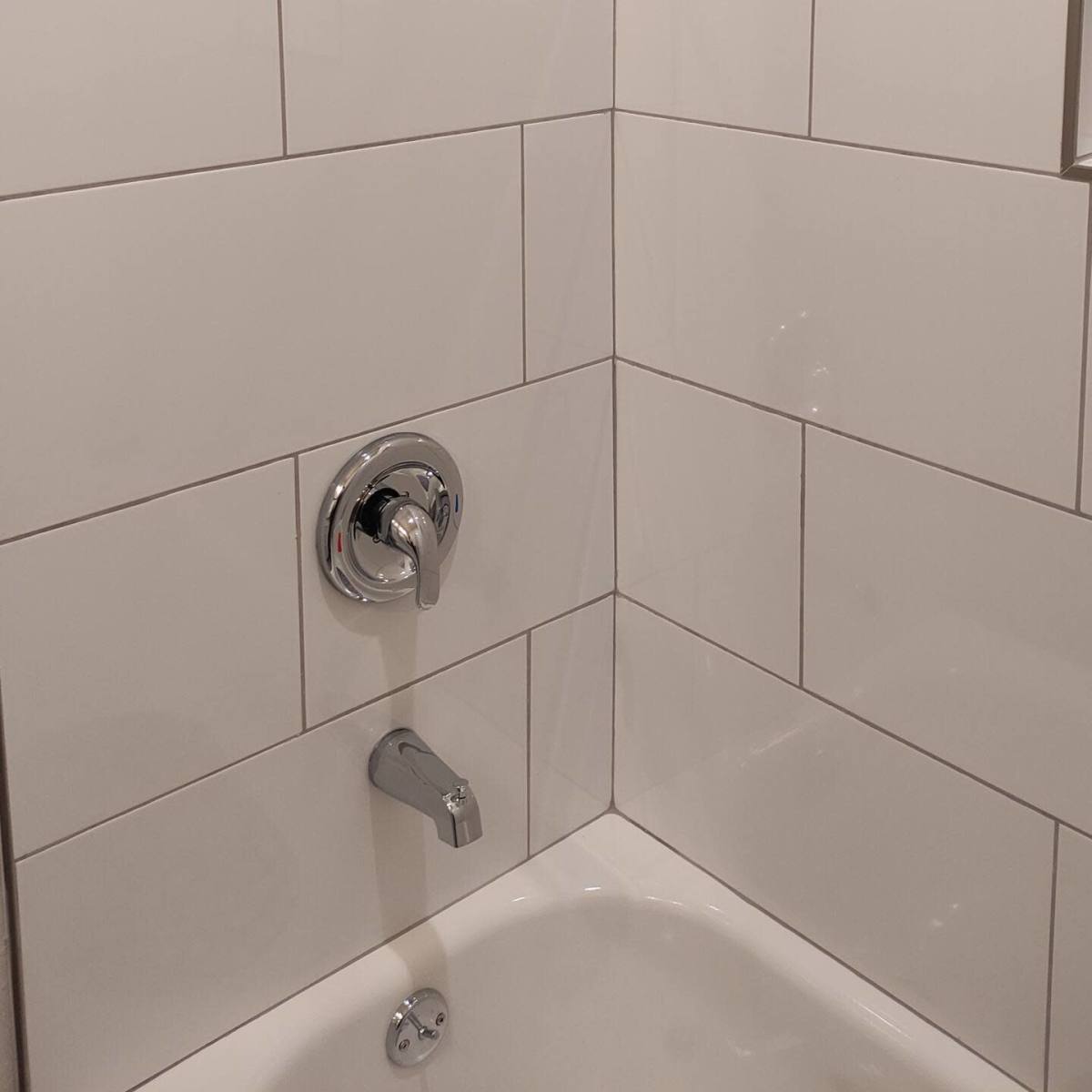 A tiled shower corner with a overhead showerhead, a wall-mounted faucet, and a water control handle.