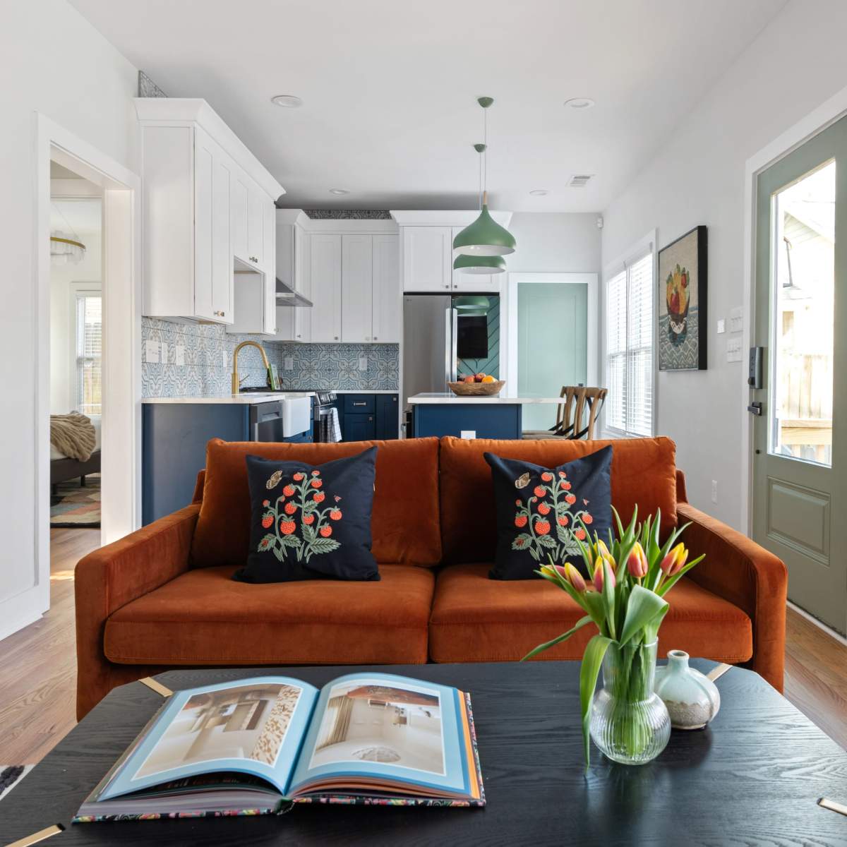 A bright living room with an orange couch adorned with floral pillows, a black coffee table, and a kitchen area visible in the background.