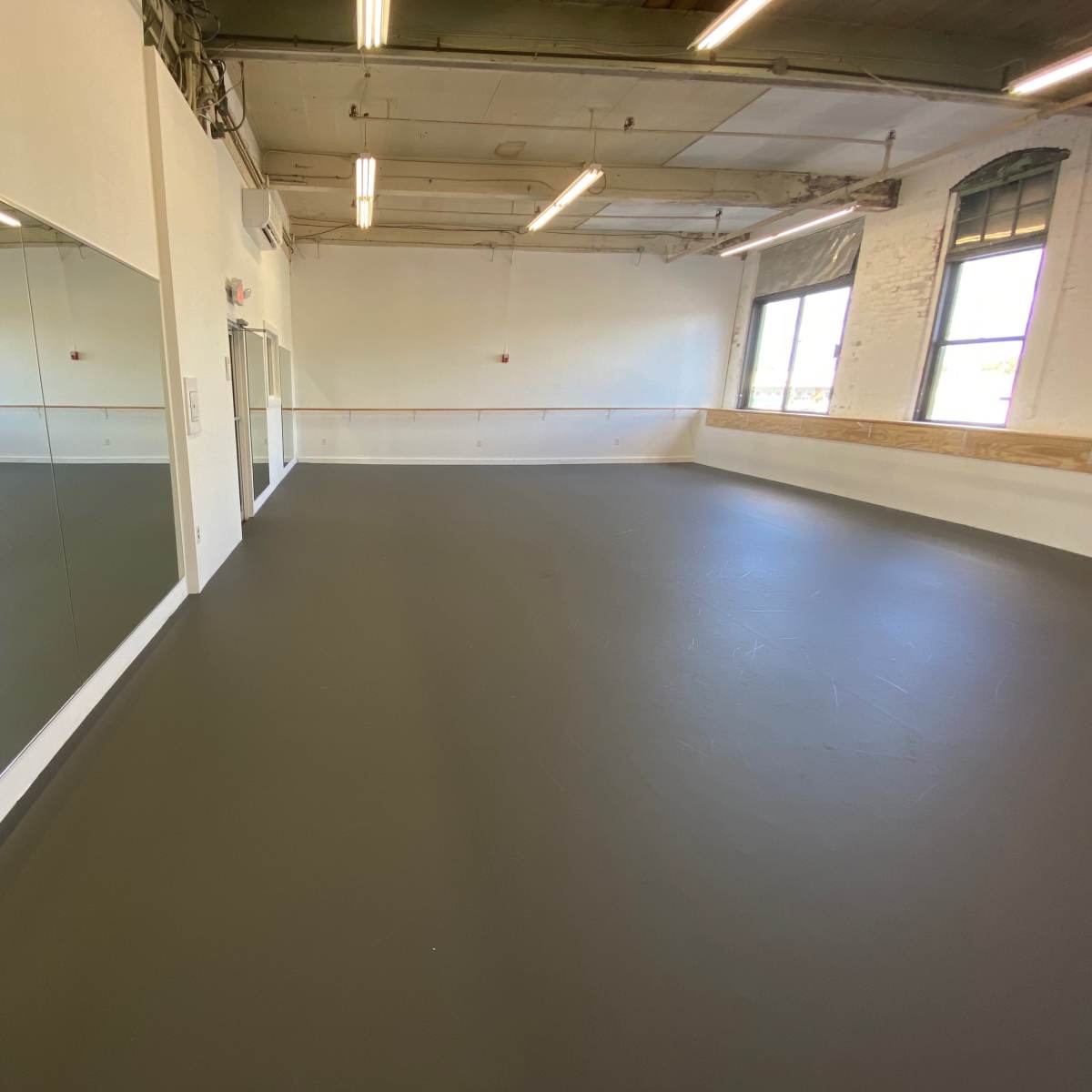 A spacious dance studio features a smooth, dark floor and large mirrors along one wall, with natural light coming through tall windows.