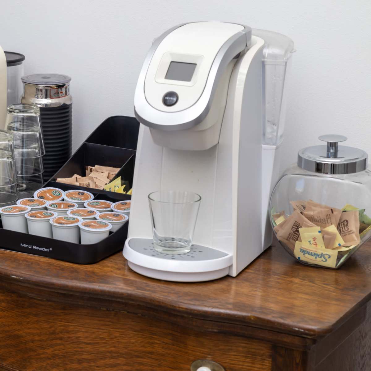 A coffee station with a cream-colored espresso machine, a white single-serve coffee maker, a glass jar of tea bags, and a tray with coffee pods arranged on a wooden dresser.
