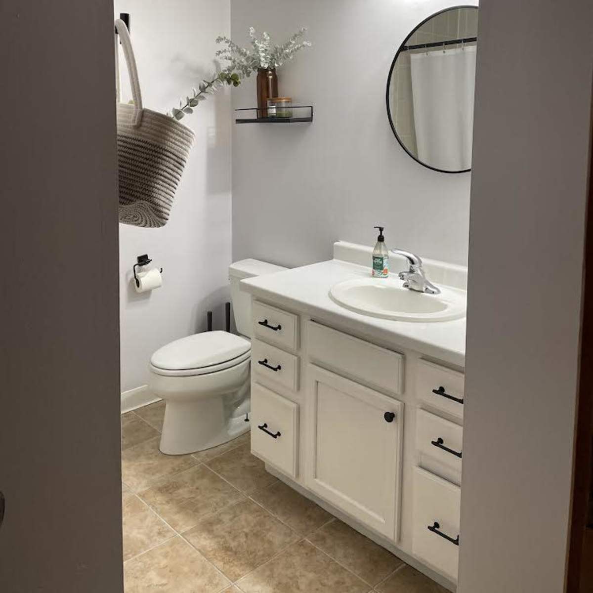 A bathroom featuring a toilet, sink with a mirror, and shelf, all against a light-colored wall and tiled floor.