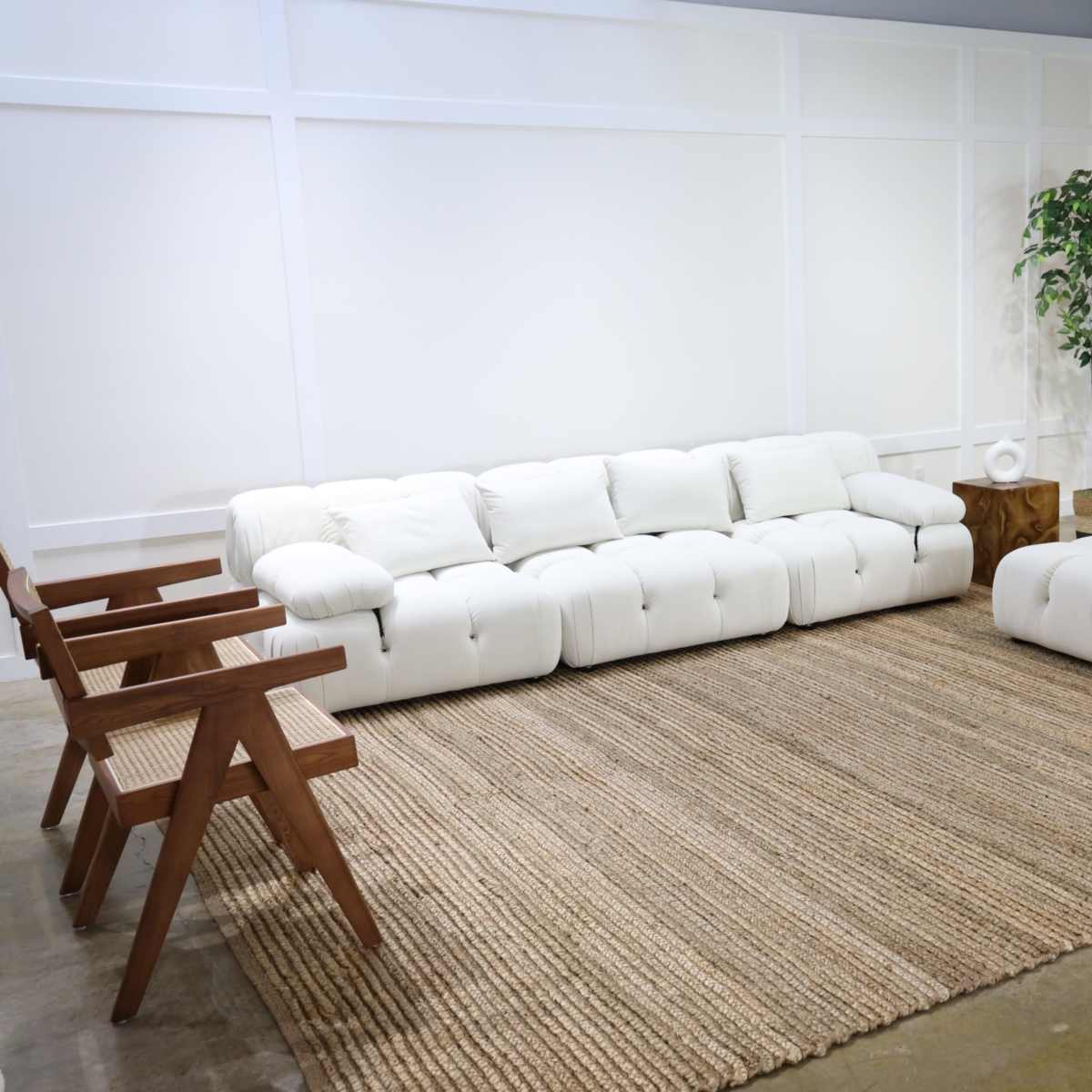 A spacious living area features a white sectional sofa, a matching ottoman, wooden chairs, and a round side table on a jute rug, with a potted plant and wooden wall accents in the background.
