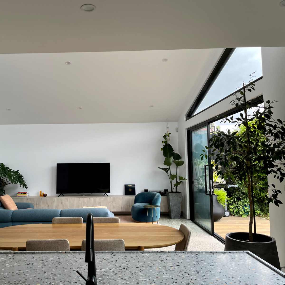 A modern living room with a sectional sofa, a large TV mounted on the wall, and plants near the windows.