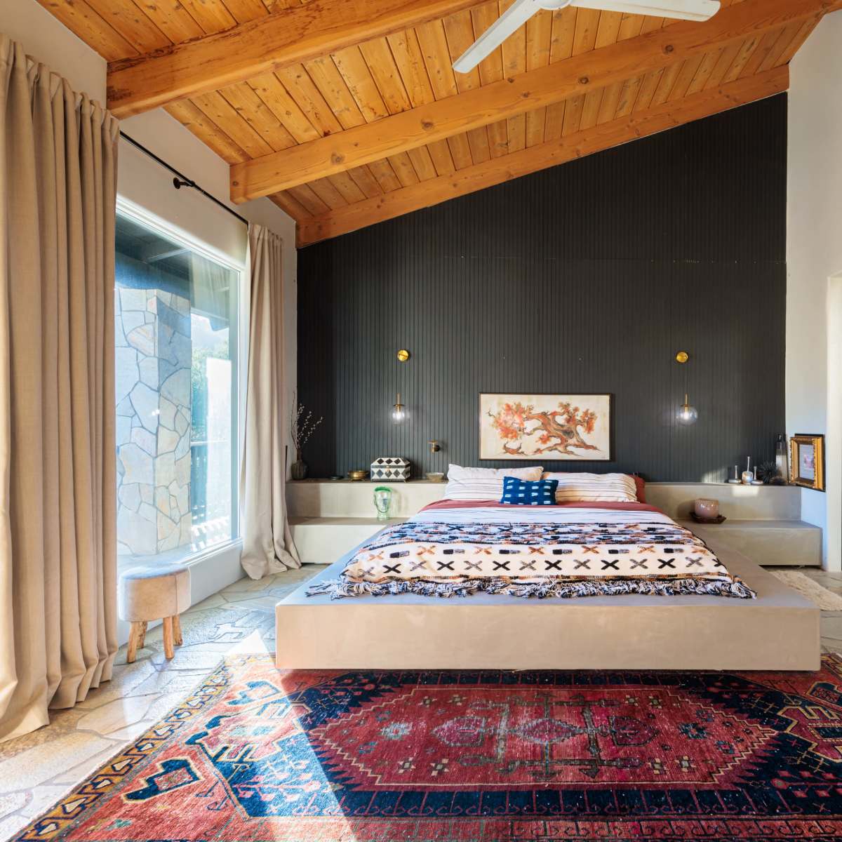 A modern bedroom with a raised bed on a platform, accented by a red area rug and wooden ceiling beams.