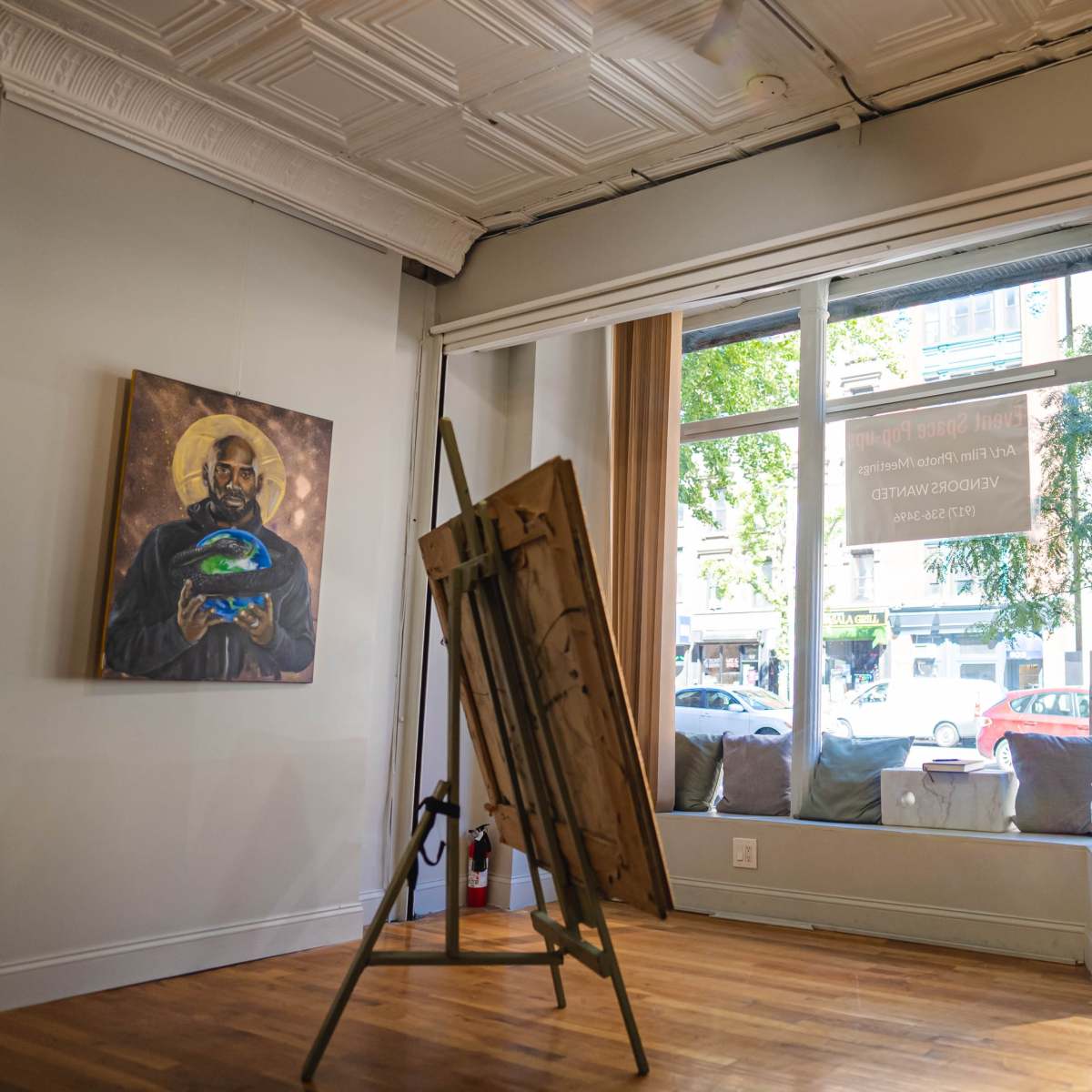 The interior of an art gallery features an easel with a painting and large windows that open to a streetscape.