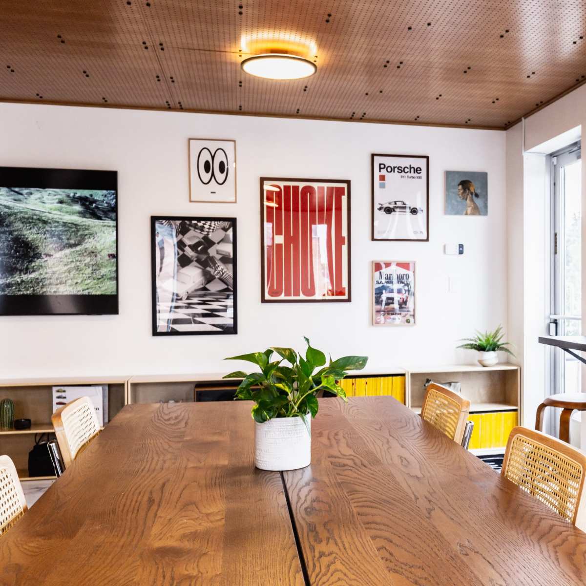 A modern workspace features a large wooden table surrounded by rattan chairs, with framed art on the walls and bar seating near large windows.