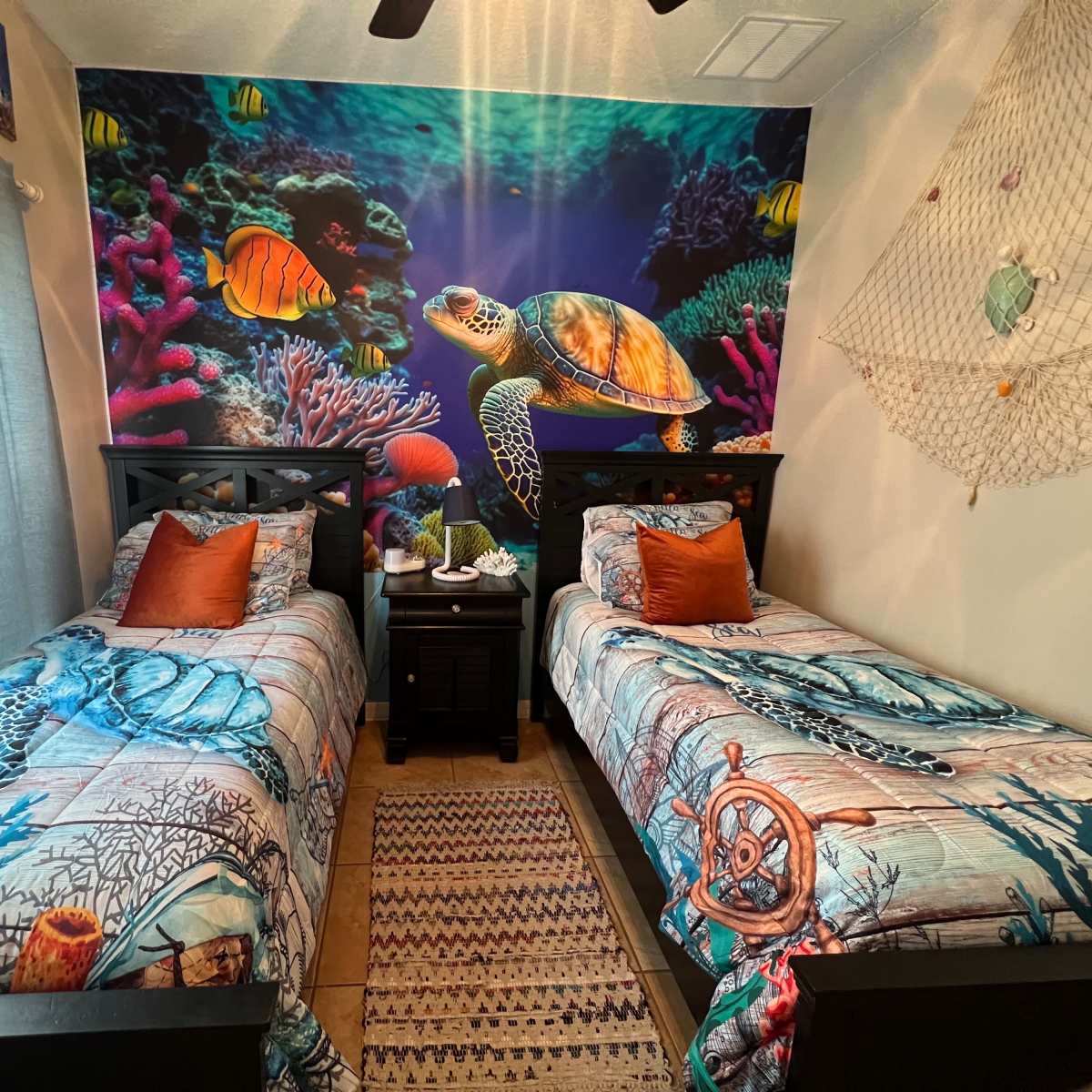 A colorful underwater-themed bedroom with two twin beds adorned with patterned bedspreads, a sea turtle mural on the wall, and decorative pillows.
