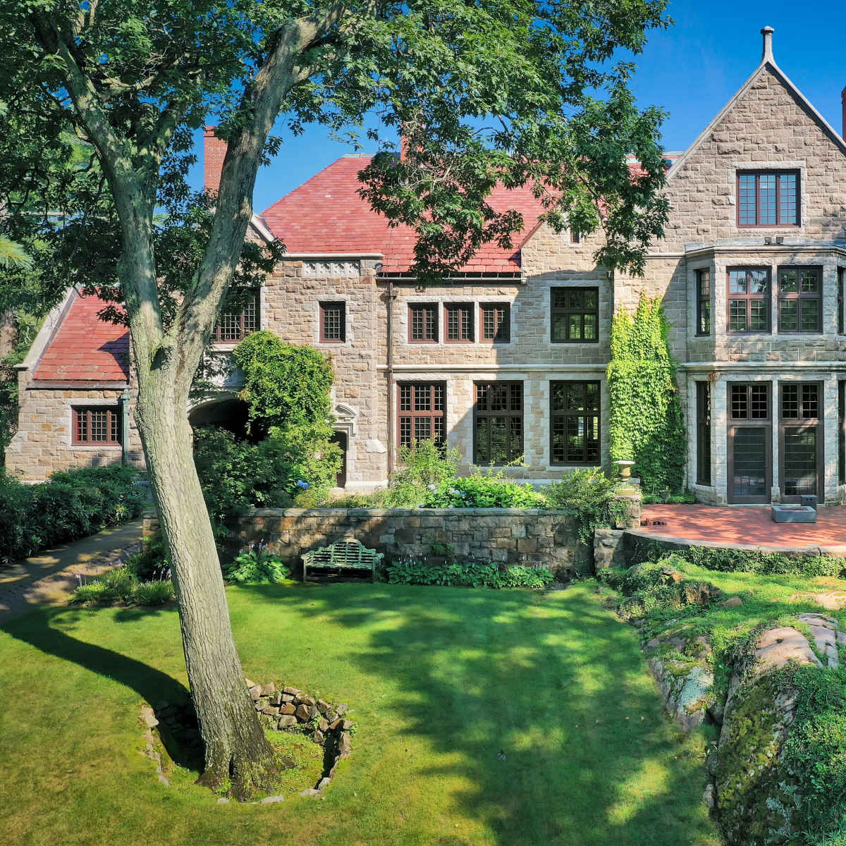 A large stone house with a red roof, surrounded by lush greenery and a manicured lawn.
