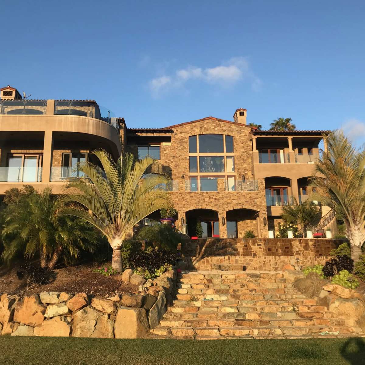 A large stone and concrete house with multiple balconies is situated on a sloped lawn, surrounded by palm trees and stone steps leading to the entrance.