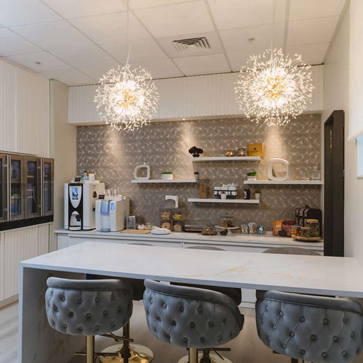A modern kitchen with a marble countertop, equipped with a coffee machine, various appliances, and decorative shelving, illuminated by two artistic light fixtures.