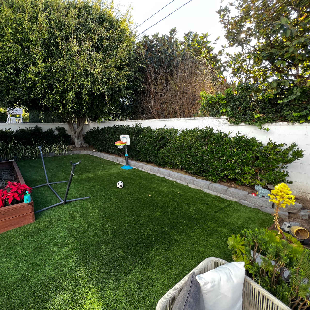 A backyard with artificial grass, a small basketball hoop, a soccer ball, and landscaped greenery along the perimeter.