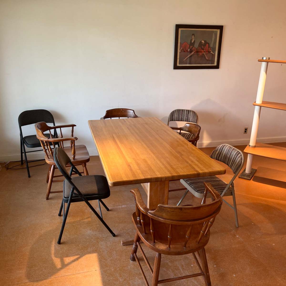 A wooden table is surrounded by several chairs in a sparsely furnished room with a window and a painting on the wall.