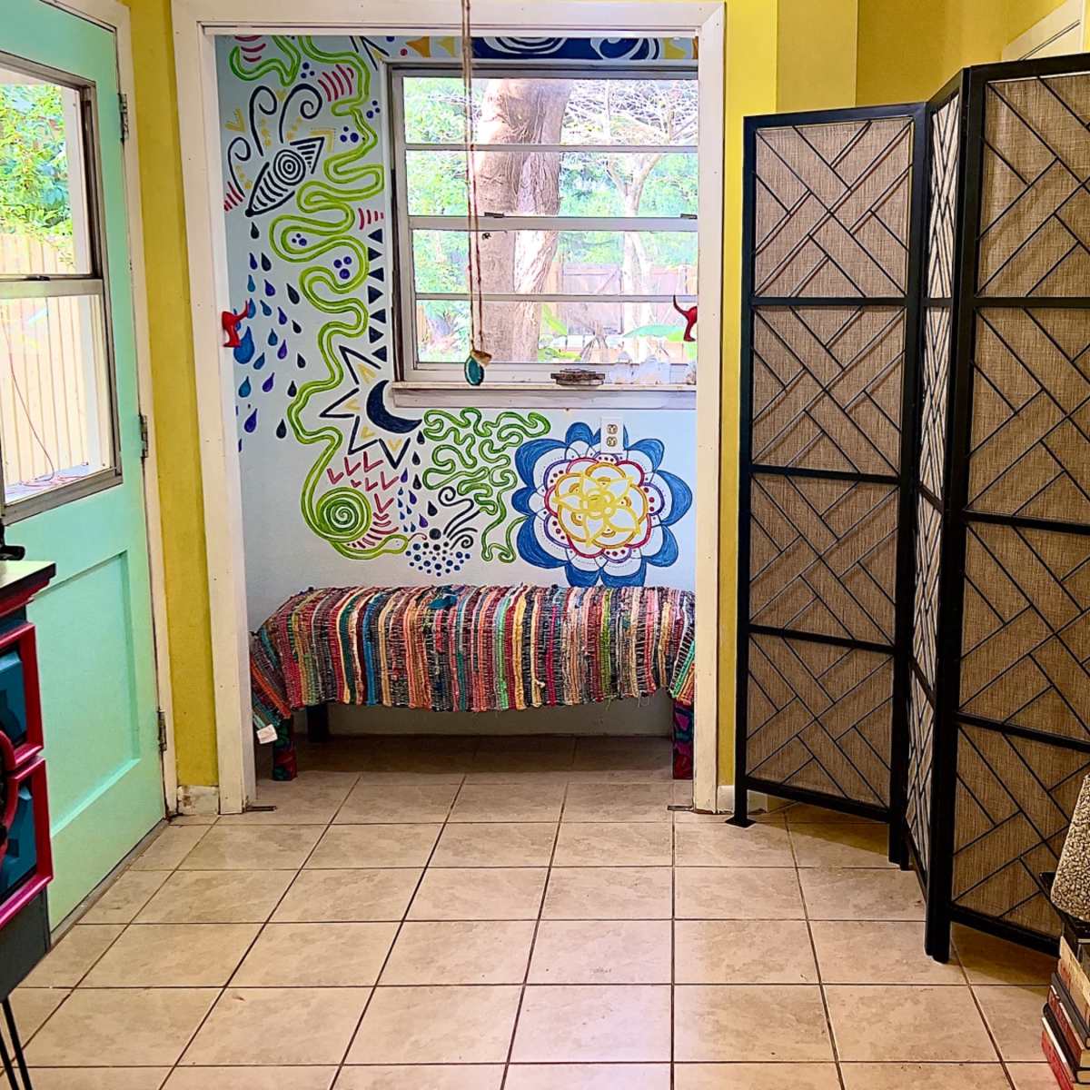 A brightly colored entryway featuring a decorative wall mural, a bench, a folding screen, and a stack of books beside a tall decorative piece.