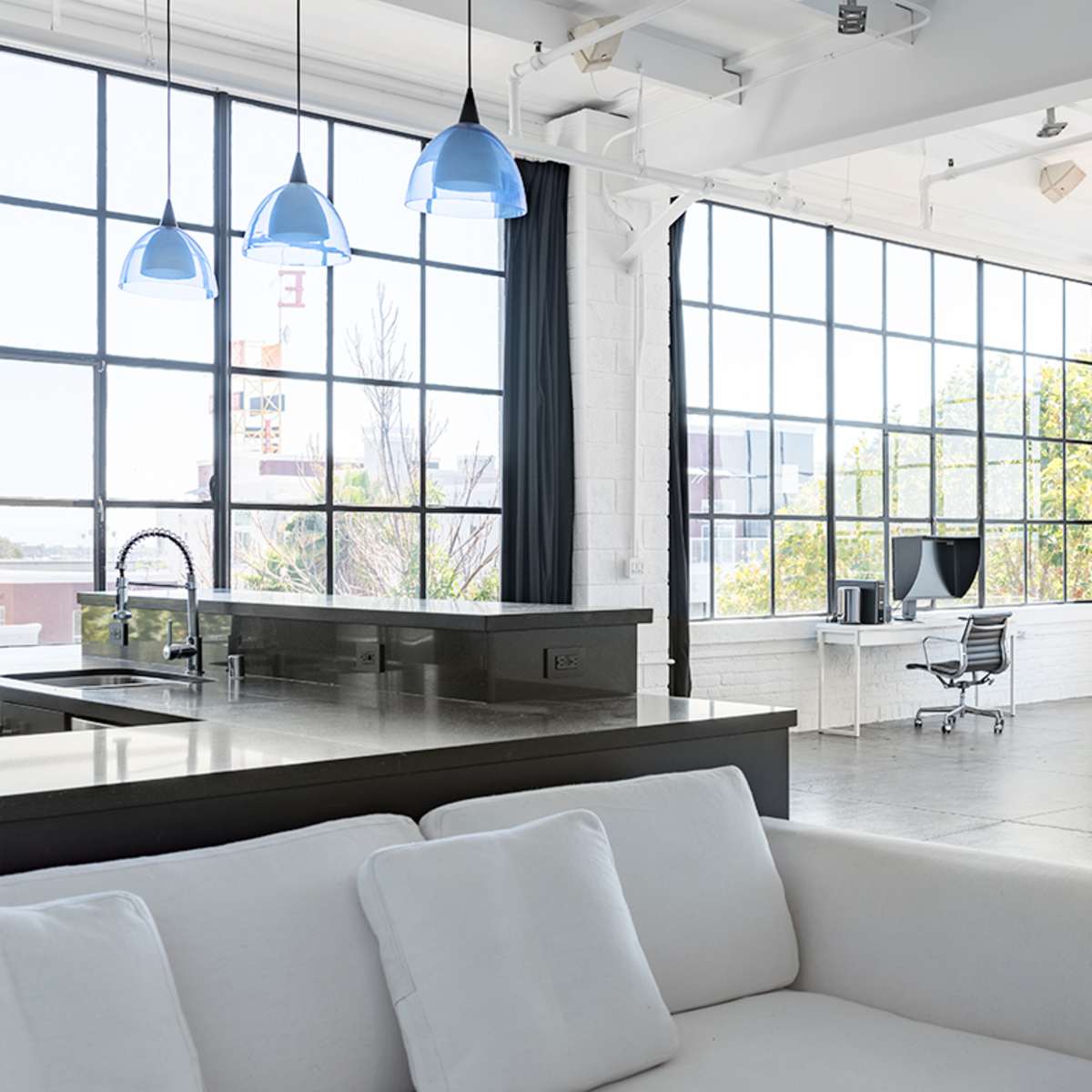 A spacious, modern loft features large windows, a minimalist kitchen island, and an organized work area with a desk and chair.