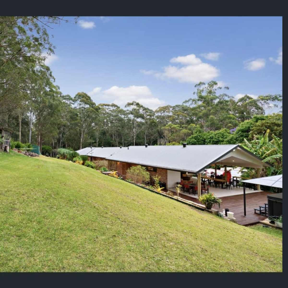 A sloping lawn leading to two modern houses nestled among tall trees and greenery.