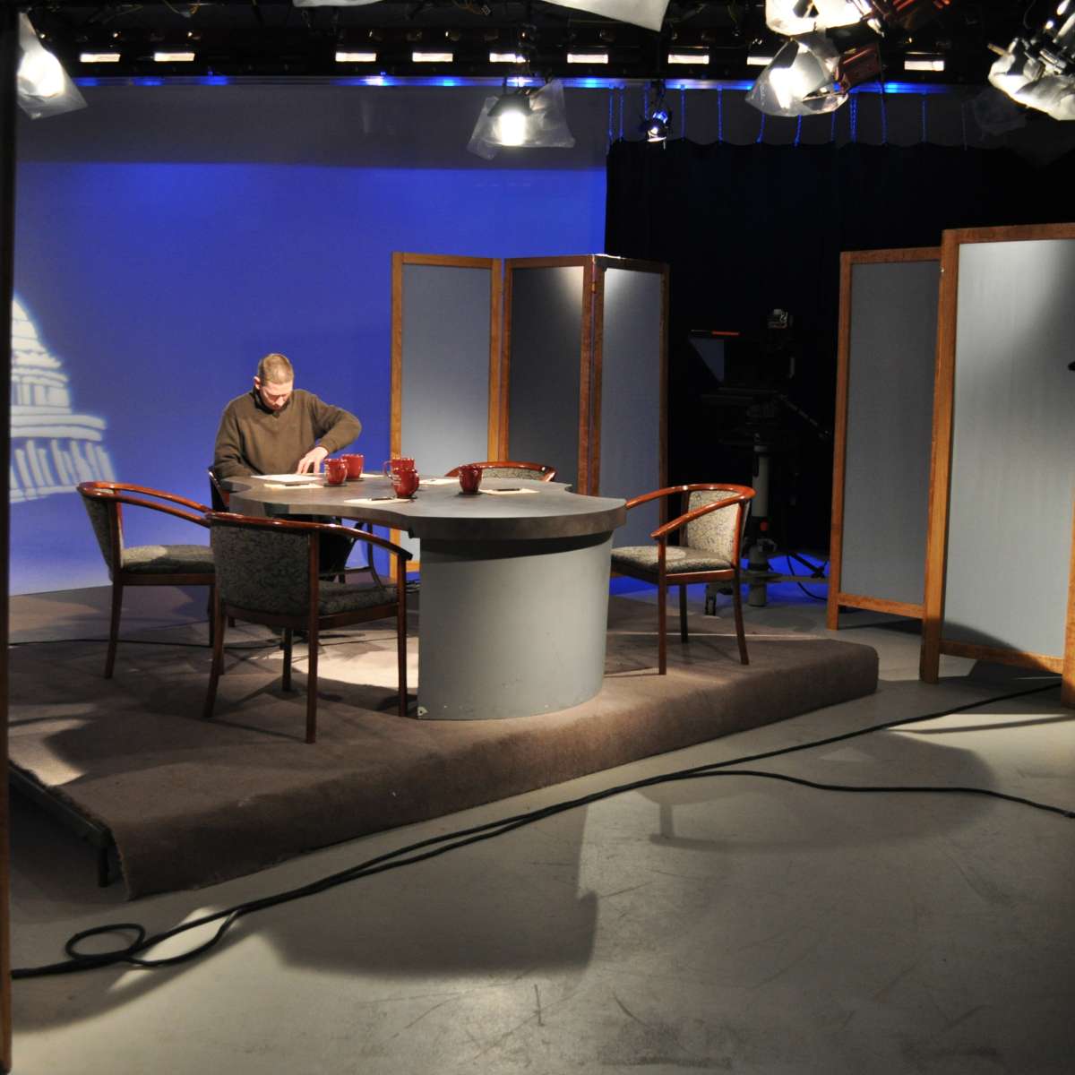 A television studio setup features a circular table with four chairs, a man sitting at the table, and backdrop screens with a faint image of a building.