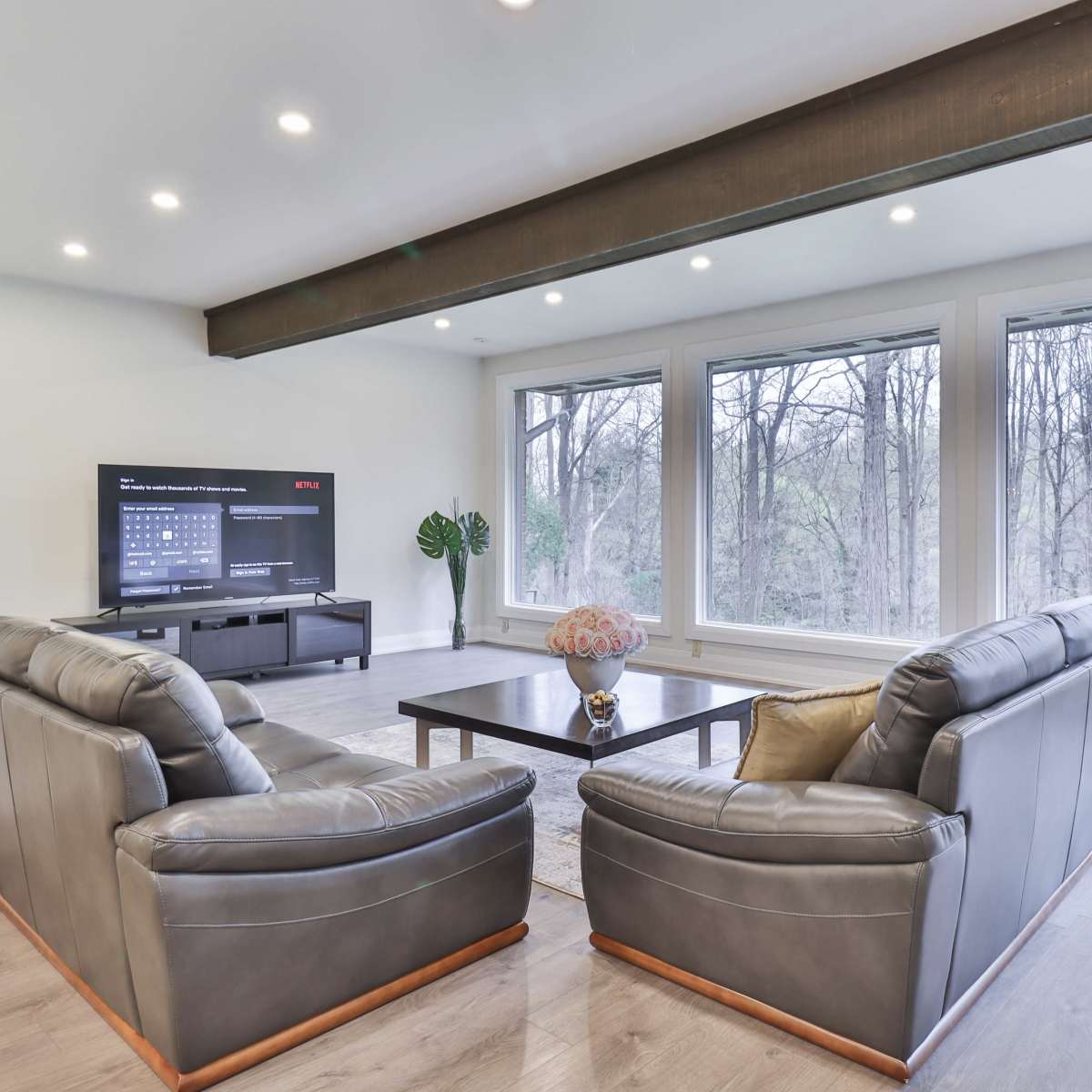The scene features a modern living room with two leather sofas facing a coffee table, a television mounted on the wall, and large windows overlooking a wooded area.