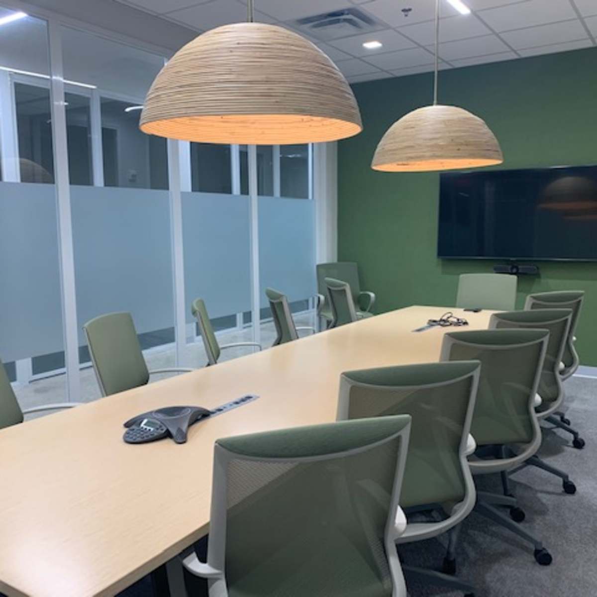 A large conference room features a long table surrounded by ergonomic chairs, with two pendant lights hanging above and a wall-mounted TV at the far end.
