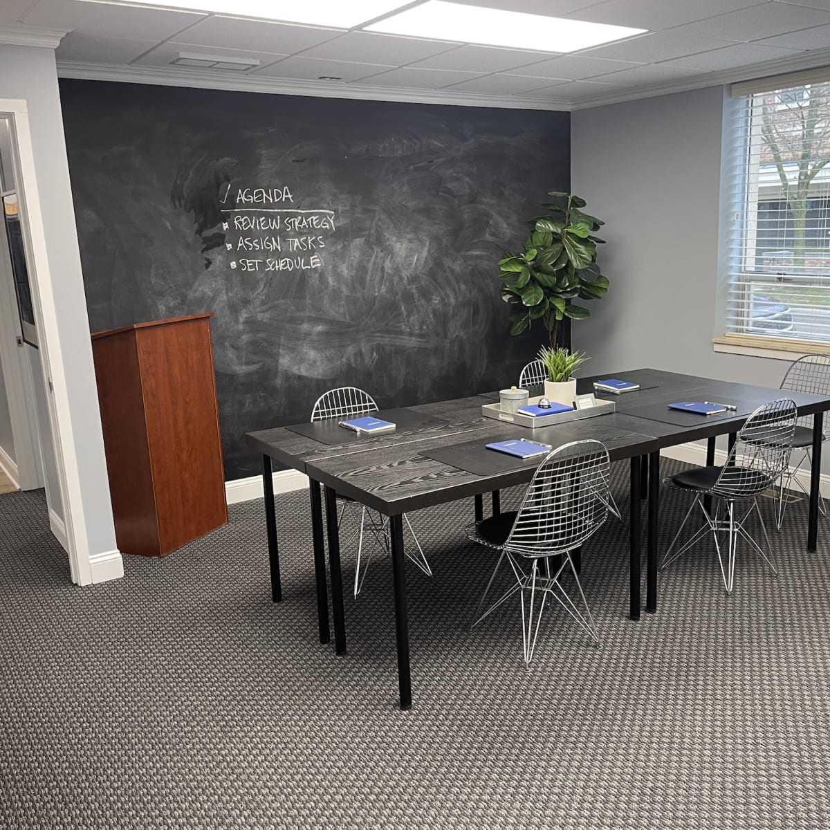 A modern conference room features a large black table surrounded by wire chairs, with a chalkboard wall listing an agenda and a potted plant in the corner.