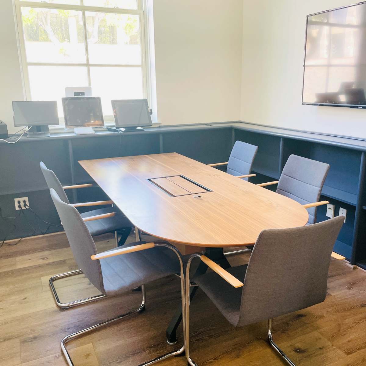 A modern conference room featuring a wooden table surrounded by five gray chairs, with a wall-mounted television and two computers on a desk in the background.