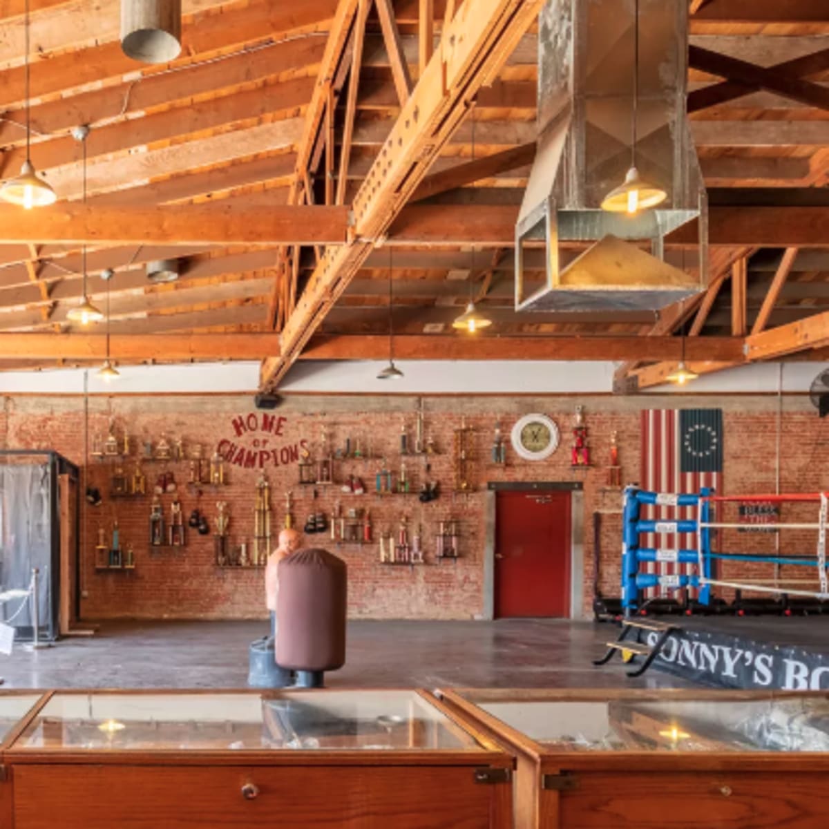 The interior of a boxing gym features exposed wooden beams, brick walls adorned with trophies, and a boxing ring in the corner.